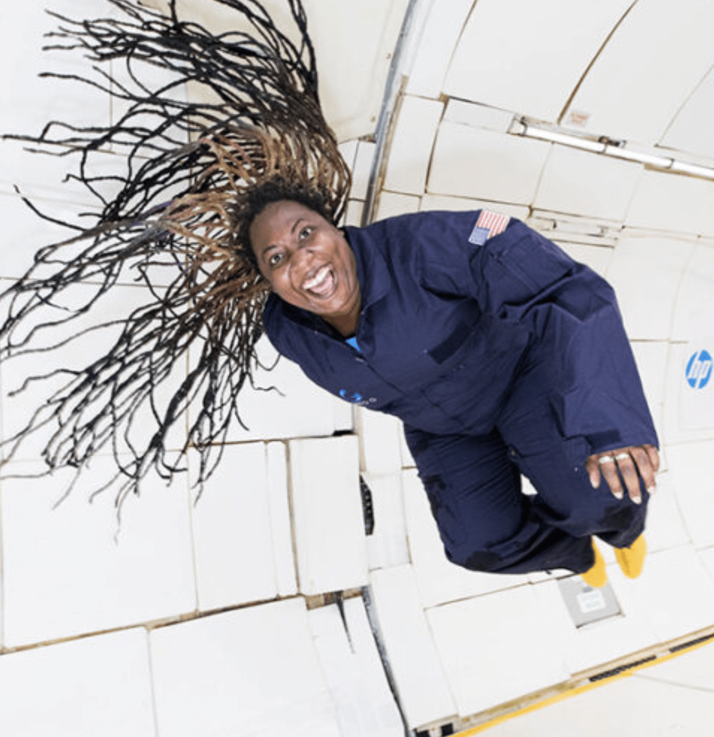 A woman wearing a navy blue spacesuit in a zero gravity environment. She has a big smile, her body is floating, and her hair is floating around her