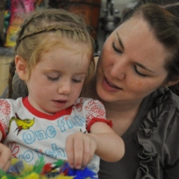 Sitting on her Mother’s lap, a blind toddler thoughtfully uses her fingers to explore a tray of multi-colored feathers