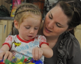 Sitting on her Mother’s lap, a blind toddler thoughtfully uses her fingers to explore a tray of multi-colored feathers