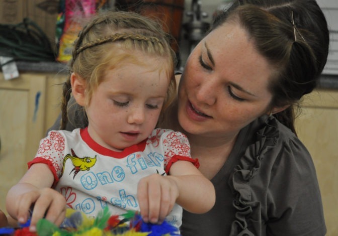 Sitting on her Mother’s lap, a blind toddler thoughtfully uses her fingers to explore a tray of multi-colored feathers