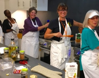 Students and instructors, all wearing white aprons, pose with cooking utensils in the LightHouse training kitchen