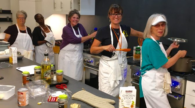 Students and instructors, all wearing white aprons, pose with cooking utensils in the LightHouse training kitchen