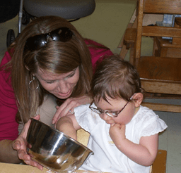 Pam sits behind a child wearing glasses who is reaching into a silver bowl Pam holds