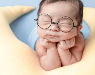 An infant, wearing glasses, who is falling asleep with his head raised, leaning on his arms on top of a moon-shaped pillow