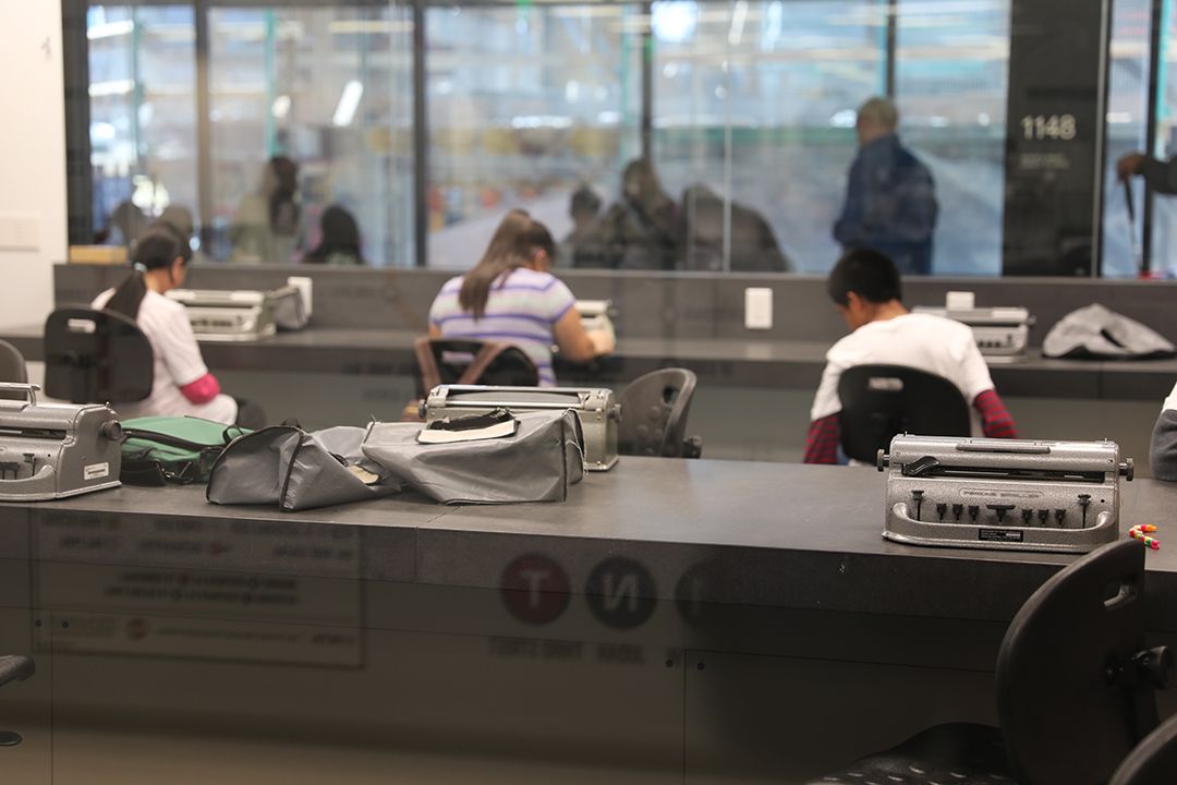 Three Perkins Braillers sit on a desk in focus in the foreground, three students working at three other Perkins Braillers are out of focus in the background
