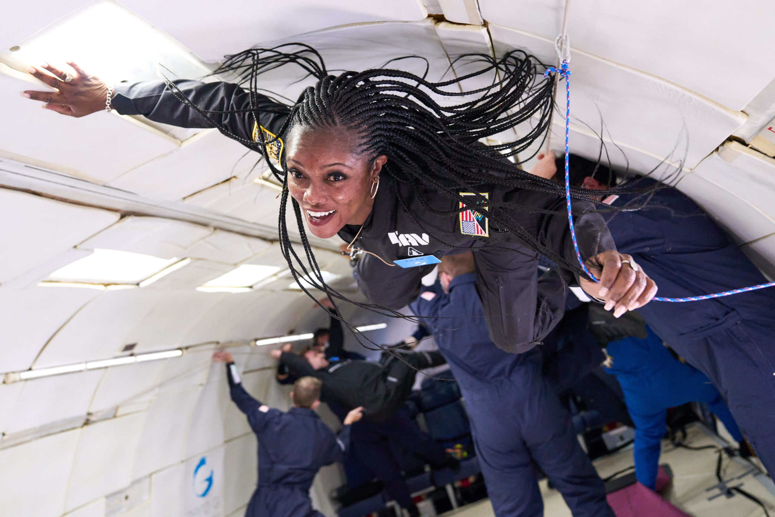 A black woman with long braids wearing a black one-piece flight suit floats suspended in mid-air inside a hollowed-out aircraft. She is loosely grasping a blue rope with her left hand