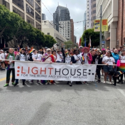 LightHouse for the Blind’s contingent in SF Pride 2022 making goofy faces at the camera. At least 15 people are holding the LightHouse banner across the road. All are wearing LightHouse’s Pride 2022 t-shirt. Participants are waving Pride flags, wearing a Pride flag as a cape, wearing a rainbow tutu, and a Guide Dog’s harness has a rainbow flag going across the bar. There are at least 30 people in the photo.