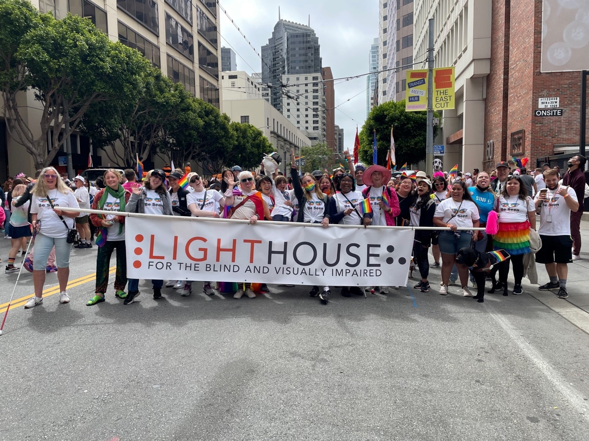 LightHouse for the Blind’s contingent in SF Pride 2022 making goofy faces at the camera. At least 15 people are holding the LightHouse banner across the road. All are wearing LightHouse’s Pride 2022 t-shirt. Participants are waving Pride flags, wearing a Pride flag as a cape, wearing a rainbow tutu, and a Guide Dog’s harness has a rainbow flag going across the bar. There are at least 30 people in the photo.