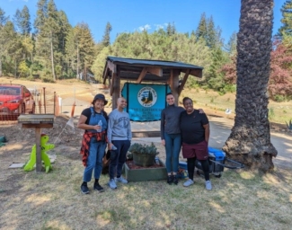 Four Verizon volunteers smile at the camera in front of the Enchanted Hills Camp sign.