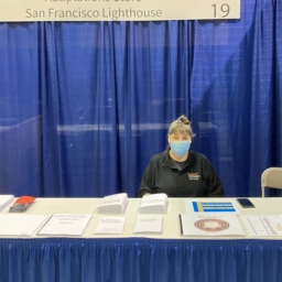 Raqi ACB 22 Adaptations store manager, Raqi Gomez, sits at the LightHouse table in the ACB exhibit hall. There are several Braille and tactile graphic products displayed on the table.