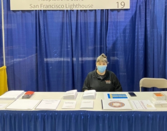 Adaptations store manager, Raqi Gomez, sits at the LightHouse table in the ACB exhibit hall. There are several Braille and tactile graphic products displayed on the table.