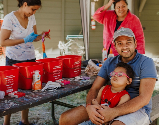 Dad holds little learner while Shruti works on the background. Another adult family member stands behind dad