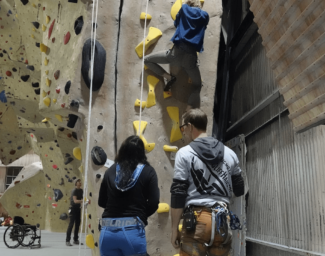 Two LightHouse students wearing rock climbing harnesses watch another student climb the rock wall in front of them