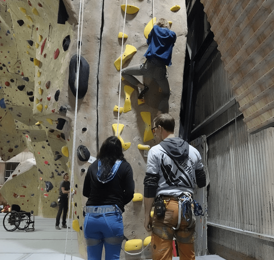 Two LightHouse students wearing rock climbing harnesses watch another student climb the rock wall in front of them