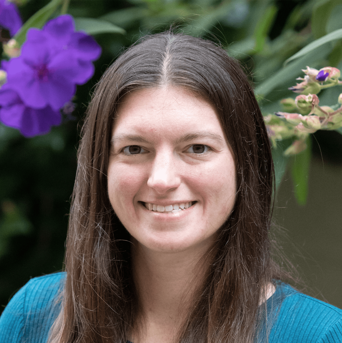 A head shot of Lauren, who has brown shoulder length hair, is wearing a teal colored top and is standing in front of green plants with purple flowers