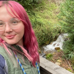 At EHC, Ellie, a young woman with pink hair, takes a selfie outdoors from the corner of a patio with a stream in the background