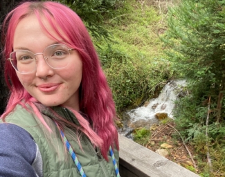 At EHC, Ellie, a young woman with pink hair, takes a selfie outdoors from the corner of a patio with a stream in the background