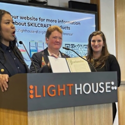 Mayor London Breed stands at a podium beside LightHouse CEO, Sharon Giovinazzo and Communications Specialist, Caitlin O’Malior
