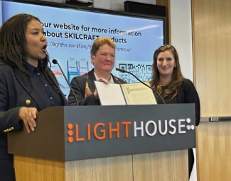 Mayor London Breed stands at a podium beside LightHouse CEO, Sharon Giovinazzo and Communications Specialist, Caitlin O’Malior