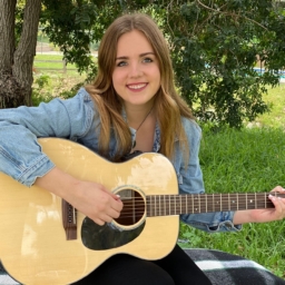 Meghan Downing sits outside on a black and white blanket and smiles while she strums a guitar.