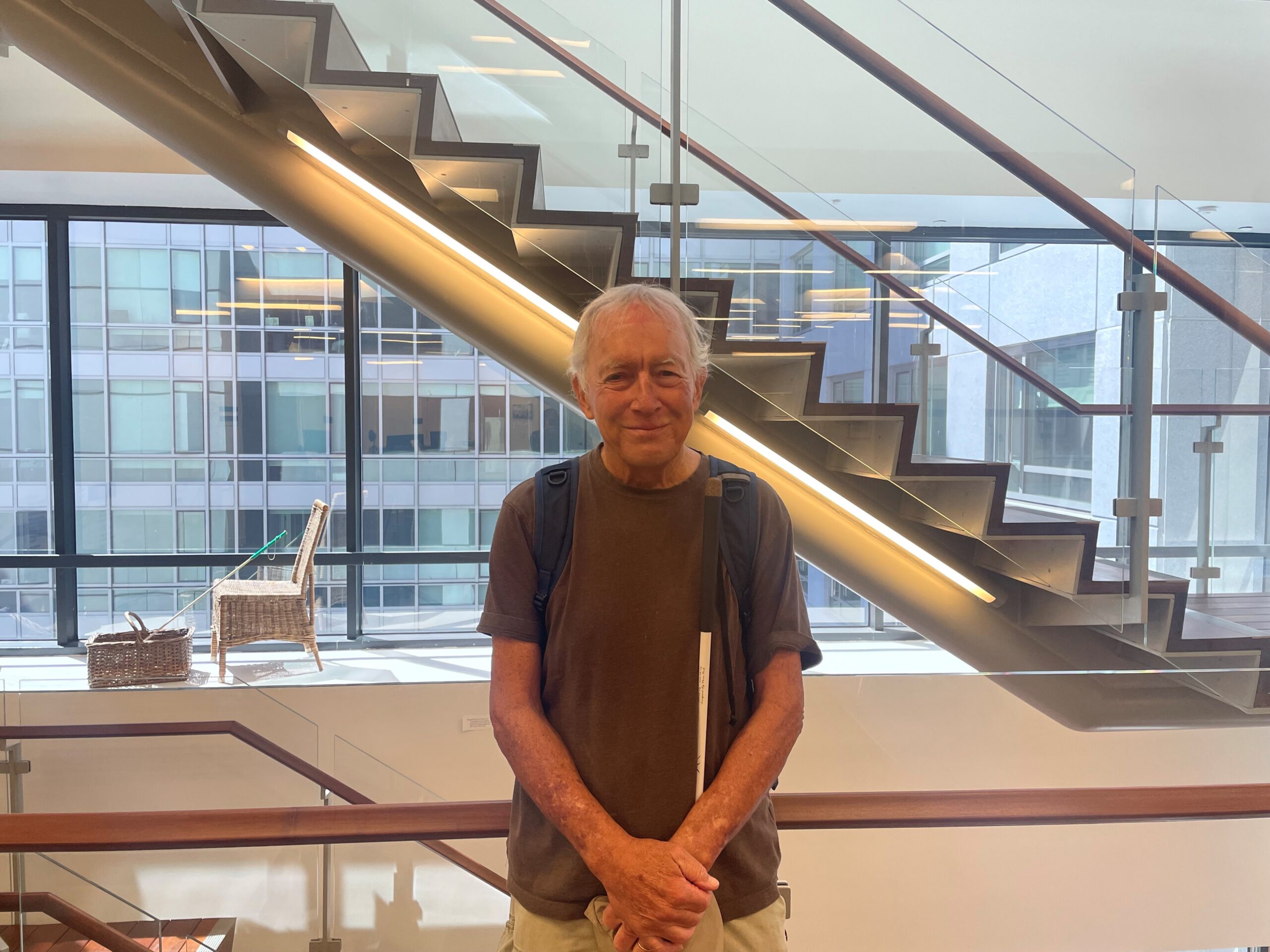 Robin Thiele, holding his white cane, stands in front of the internal staircase on the 10th floor of LightHouse headquarters