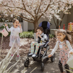 1. Three children smile holding Easter baskets, the child in the middle is a wheelchair user