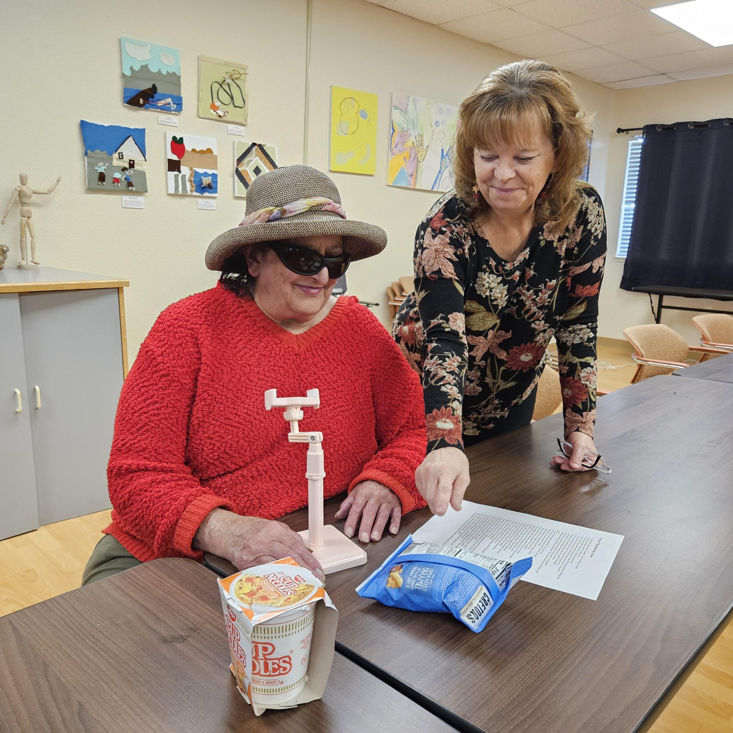 Cathy, right, stands and smiles as she watches a client seated with their hand on a phone stand that can be used to take photos for text recognition. On the table are a piece of paper, a bag of chips and a Cup of Noodles.