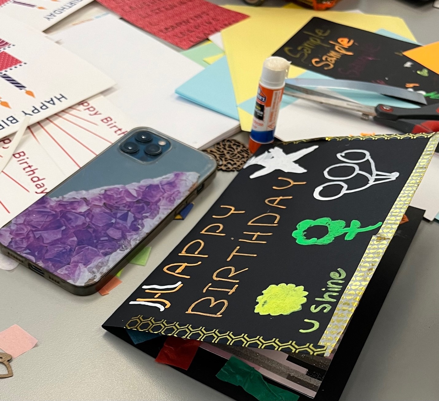 A cell phone and various card and letter writing supplies on a table, including a black card with washi tape edges and “Happy Birthday U Shine” and flowers drawn on the front with high contrast paint pens.