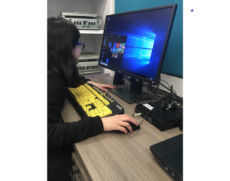 A student sits at a desktop computer with a large print keyboard with black on yellow keys