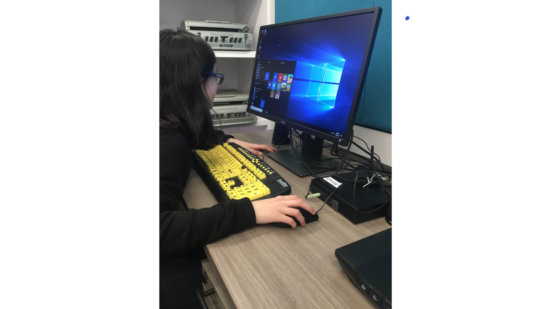 A student sits at a desktop computer with a large print keyboard with black on yellow keys