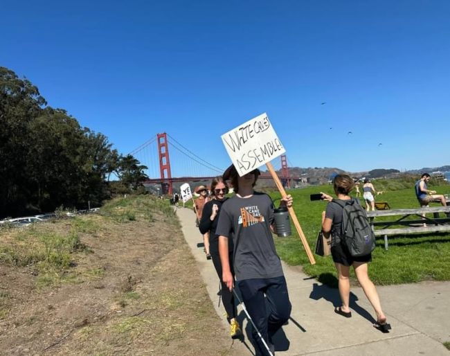 Marchers walk down the dirt path from the Golden gate Bridge to the Chrissy Field after-party. One sign reads, “White Canes Assemble!”