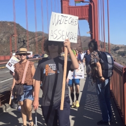White Cane Day marchers cross the Golden Gate Bridge. Center, a white cane user carries a sign that reads “White Canes Assemble”.