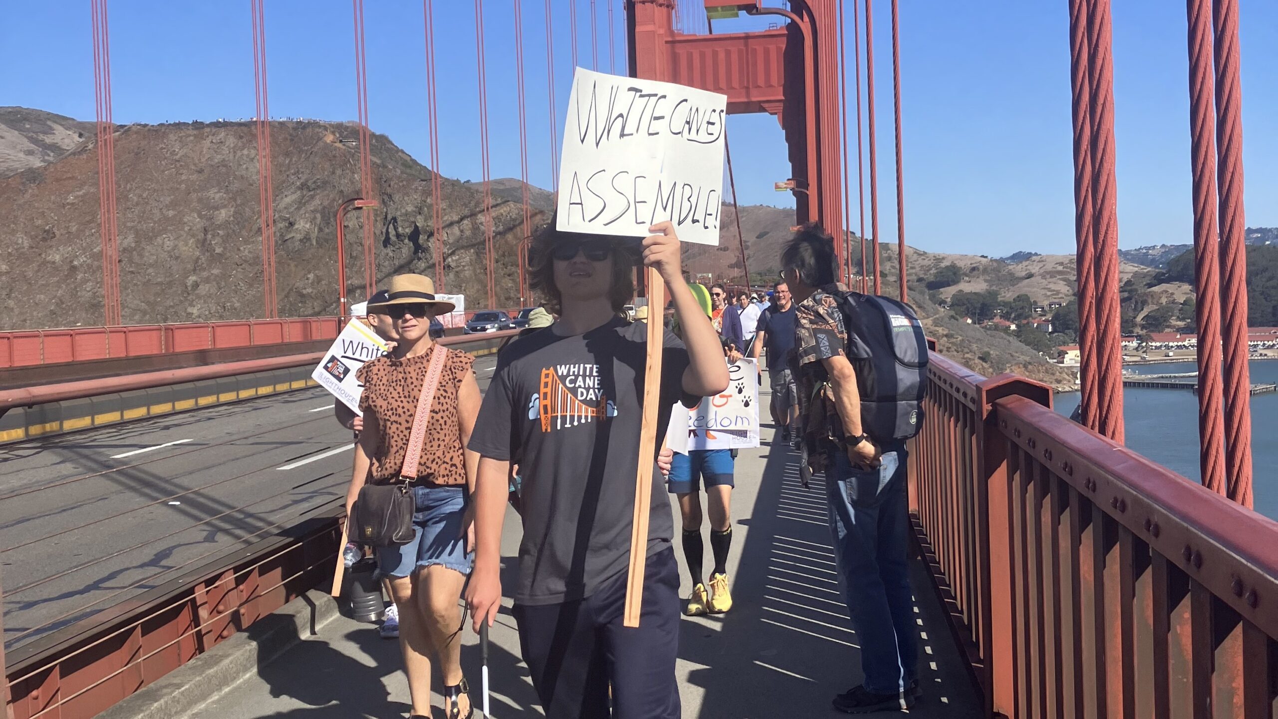 White Cane Day marchers cross the Golden Gate Bridge. Center, a white cane user carries a sign that reads “White Canes Assemble”.