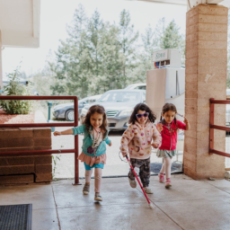 Three Little Learner campers, one using a white cane, run together past the dining hall at Enchanted Hills Camp.