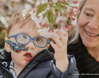 A young child, wearing glasses, reaches out to touch some flowers hanging from a plant as an adult looks on.