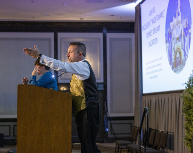 Auctioneer Greg Quiroga wears and gold vest and stands at the podium talking excitedly with his hands in the air