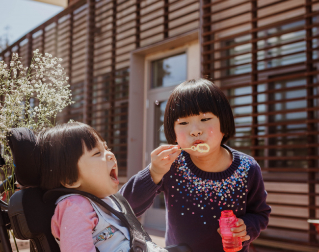 A little girl who is a wheelchair user expresses delight as her sister, who is standing next to her, blows bubbles