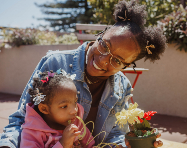 A mother cuddles with her daughter who is playing with a rainbow slinky toy. The mother is smiling and holding onto a silk flower planted in a pot.