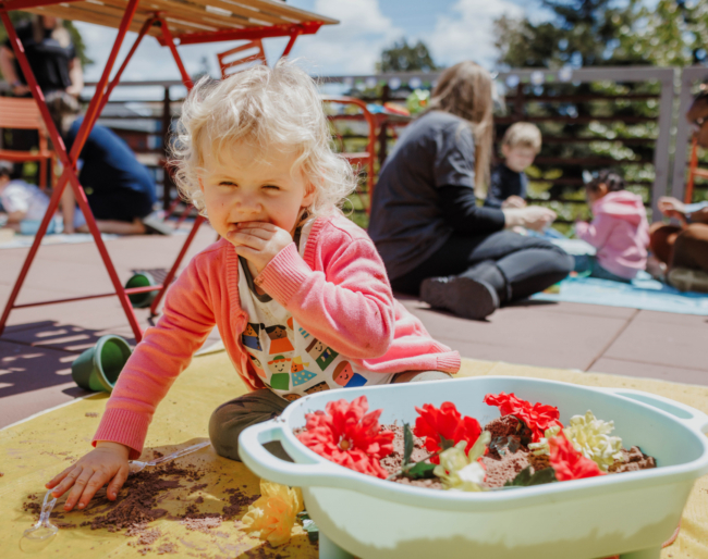 A toddler sits next to a plastic bin of silk flowers and raises her fingers to taste the edible soil